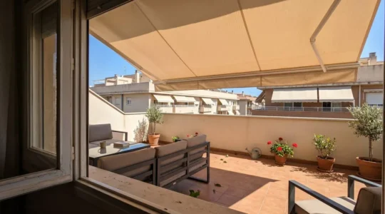 Vista desde interior vivienda hacia terraza residencial con toldo beige desplegado proyectando sombra definida sobre suelo de baldosas, mobiliario parcialmente visible en zona sombreada